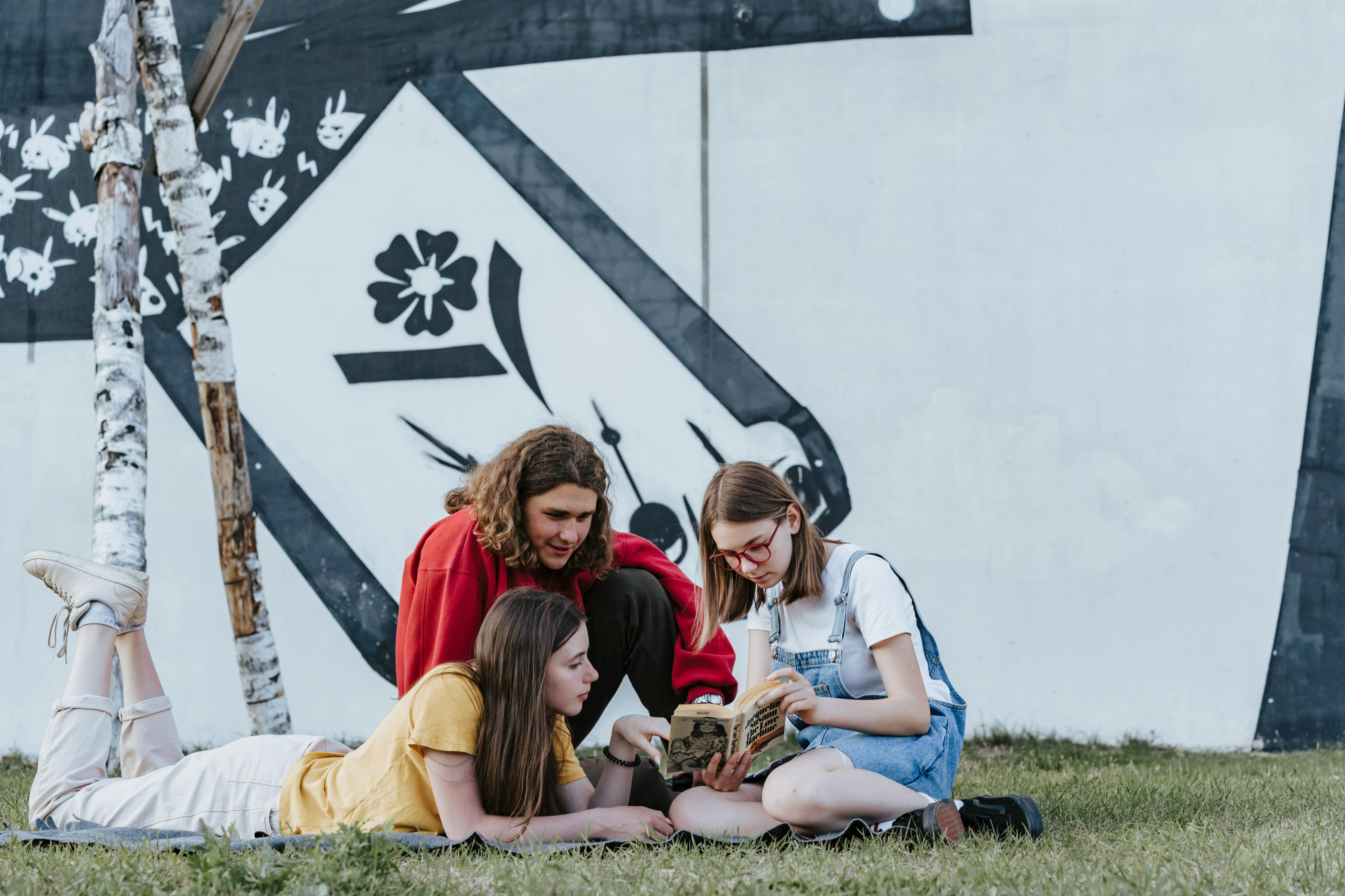 A group of students gathered at the park sitting on the grass reading a book