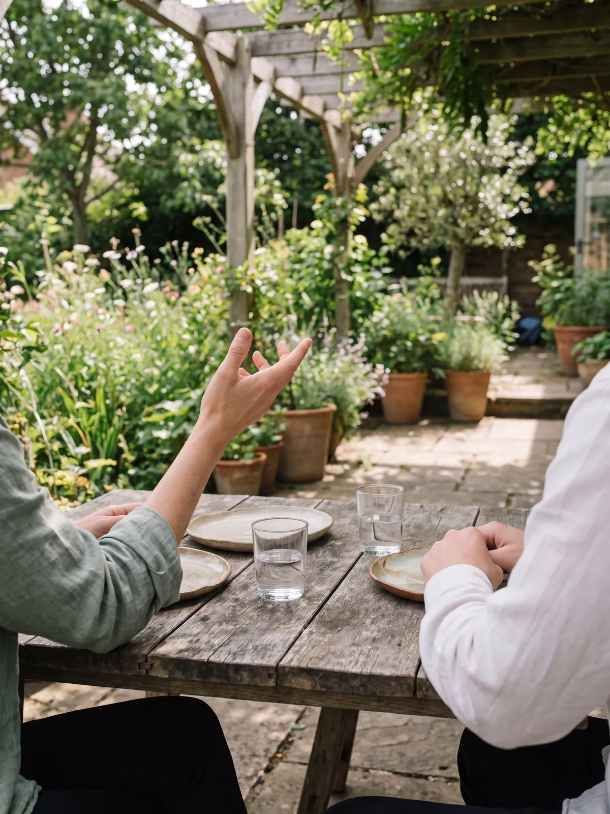 Two people talking across a weathered wooden table in a leafy garden courtyard